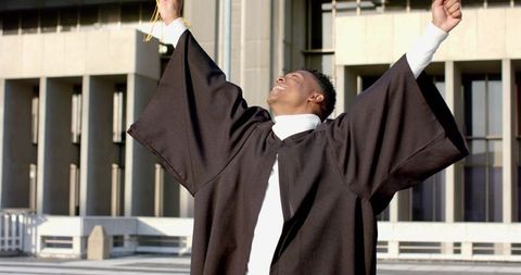 African american graduate celebrating on campus terrace raising arms with gold tassel