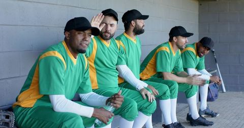 Dugout teammates wearing green and yellow baseball uniforms sitting and talking on bench