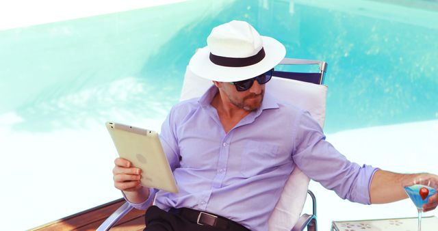 Man in a light blue shirt and white hat sitting poolside while reading from a tablet and holding a drink. Sunglasses add to the relaxed vibe. Perfect for themes related to summer, relaxation, luxury, vacations, leisure activities, technology use outdoors, and lifestyle content.