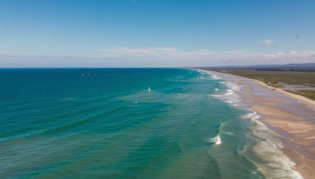 Aerial coastline curving along wide sandy shore with turquoise surf and distant sailboats