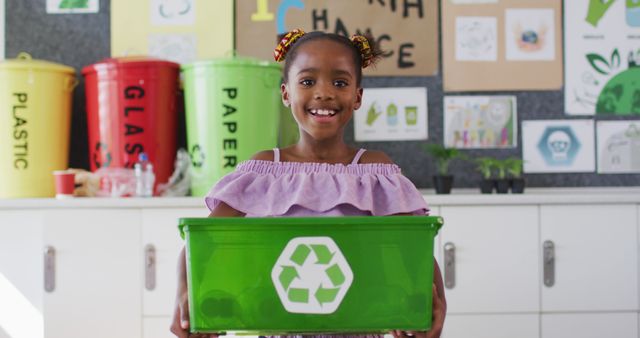 Young African American girl smiling and holding a green recycle bin in an educational setting focused on recycling and environmental awareness. This is useful for themes on sustainability, children's education, environmental care, and community awareness initiatives, emphasizing the importance of teaching ecological habits to younger generations.