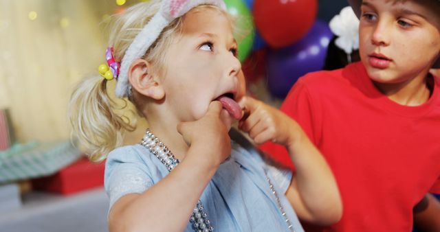 Young children playfully making funny faces during a festive birthday party with colorful balloons in background. Perfect for use in advertisements, family blogs, children's event invitations or educational materials that highlight childhood fun and playfulness.