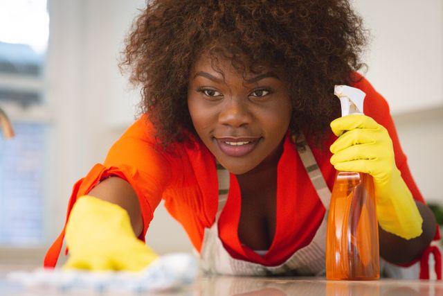 African American woman wearing yellow gloves and an apron, cleaning a kitchen counter with a spray bottle and cloth. This image can be used for articles or advertisements related to household chores, cleaning products, hygiene, and domestic work. It is also suitable for illustrating topics on home maintenance, cleanliness, and sanitation.