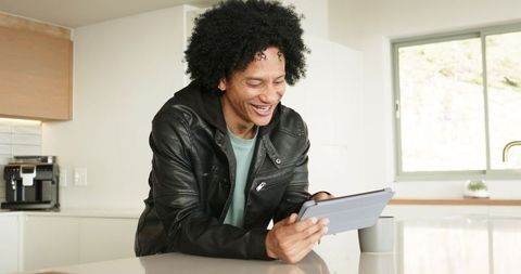 Mid-adult African-American man leaning on modern sunlit kitchen counter using tablet