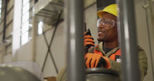 Construction worker using walkie talkie in warehouse. He wears safety gear including a hard hat, protective eyewear, and orange gloves while communicating effectively with others. Useful for depicting industrial work, safety protocols, and communication in a professional environment.
