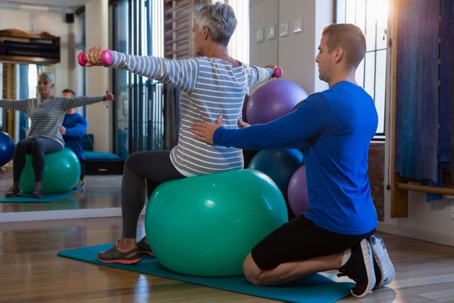 Physiotherapist assisting senior woman on exercise ball and dumbbells at clinic