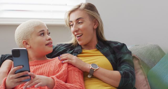 A teenager is using a smartphone while seated on a couch next to an adult who is smiling at them. This scene is perfect for advertisements or articles about family time, modern technology in everyday life, or positive relationships between teens and adults. It conveys a sense of bonding, comfortable setting, and modern family dynamics.