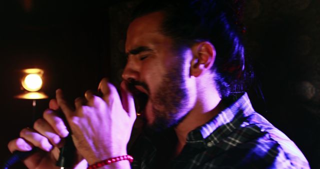 A young Latino male passionately sings into a microphone, illuminated by dramatic stage lighting, with copy space. His intense expression and dynamic pose suggest a live music performance or a moment of artistic expression.