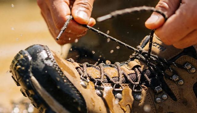 Tying wet hiking boot laces at muddy puddle, hands tightening rugged outdoor footwear