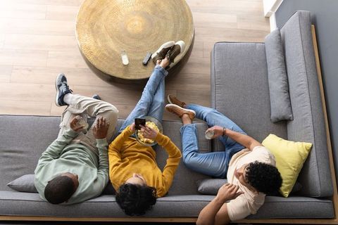 Diverse friends relaxing on gray sofa sharing snacks, drinks and scrolling smartphone