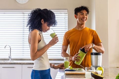 Diverse couple tasting fresh green smoothie after blending in bright home kitchen