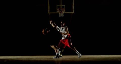 Two basketball players battling under spotlight at gym hoop driving to rim with intensity