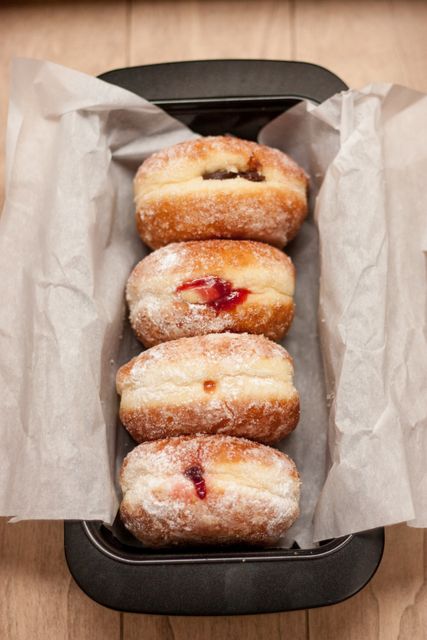 Image shows freshly baked jelly doughnuts lined up in a baking tray covered with parchment paper on a wooden surface. Ideal for use in food blogs, recipe books, bakery advertising, or social media posts focused on homemade desserts and baking.