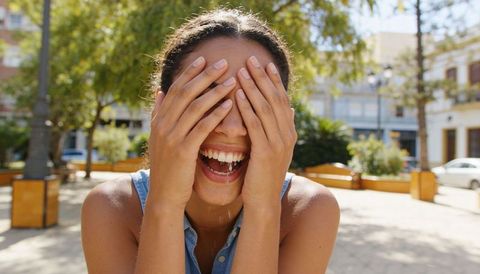 Joyful woman laughing and covering eyes with hands in sunlit urban plaza