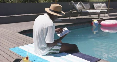African man reading paperback by pool wearing straw hat enjoying leisurely summer escape