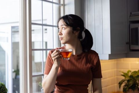 Asian woman sipping tea and gazing through window in modern kitchen, contemplative morning light