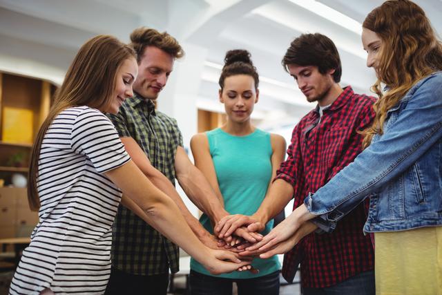 This image portrays a group of young professionals standing in a circle with their hands stacked together, symbolizing teamwork and unity. With a casual office backdrop, it showcases team collaboration and the supportive work culture in modern businesses. Ideal for use in articles, presentations, and promotional materials about team building, company culture, employee motivation, and collaborative work environments.