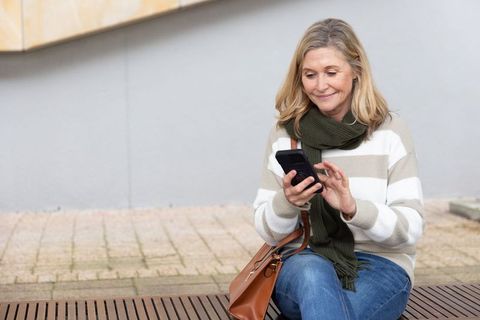 Mature woman using smartphone on bench wearing striped sweater, scarf and brown handbag
