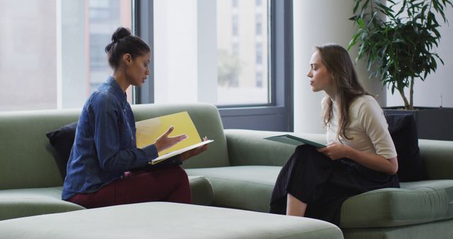Two women are sitting in a modern office space, engaged in a discussion while holding business documents. One woman is gesturing as she speaks, while the other is listening attentively. The setting includes a contemporary office environment with large windows and a potted plant. This image can be used to illustrate themes of professional collaboration, teamwork, diversity in the workplace, business meetings, and modern office settings.