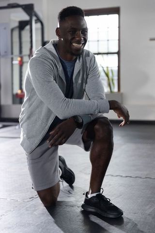 African american man kneeling in gym smiling after workout wearing hoodie and smartwatch