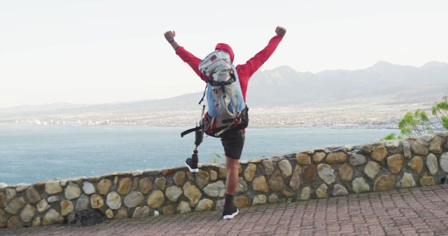 Rear view of biracial man with prosthetic leg trekking, raising arms in victory looking out to sea. Long distance walking, fitness, challenge, disability, nature and healthy outdoor lifestyle.