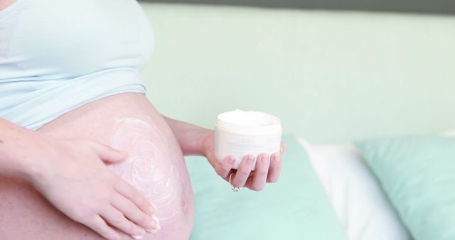 A pregnant Caucasian woman is applying cream to her belly to moisturize and care for her skin, with copy space. Pregnancy skincare routines often include such products to help reduce the appearance of stretch marks and maintain skin elasticity.