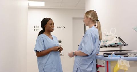 Two nurses discussing patient care in radiology corridor with mobile equipment