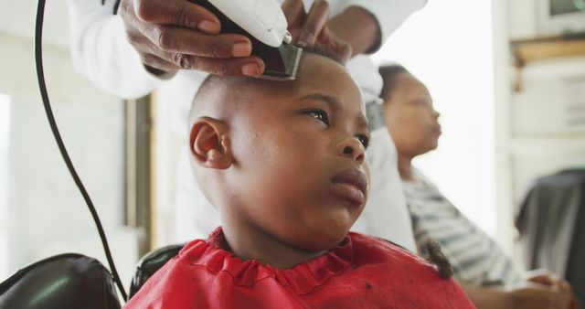 Young boy receiving a haircut at a barbershop, facing the camera with an intense look, while an adult behind him focuses on their own task. Ideal for topics related to grooming, personal care, childhood, barbershop culture, and male grooming services.