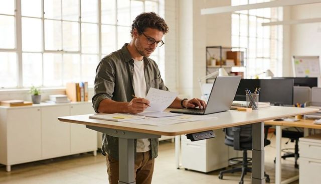 Man reviewing documents at adjustable standing desk in bright open office with laptop