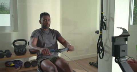Man rowing on ergometer in home gym with kettlebell and trx for functional fitness