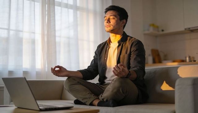 Young man meditating on sofa with laptop during golden hour practicing mindfulness at home