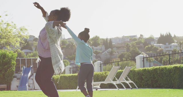 Two African American females bonding by dancing outdoors in a sunny park with green grass and lounge chairs visible. Ideal for themes related to family, outdoor activities, joy, happiness, and carefree childhood moments. Suitable for use in parenting blogs, lifestyle magazines, advertisements, and social media post. The image reflects energy and positive familial interactions.