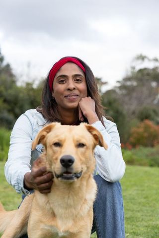 Indian woman petting golden dog in park wearing denim jacket and red headband smiling