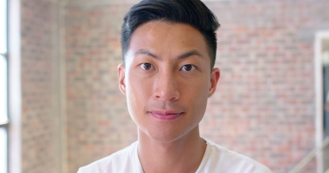Young Asian man with short hair smiling confidently at the camera. Behind him is a brick wall with part of a window in a modern indoor setting. Ideal for use in articles or marketing materials promoting confidence, young professionals, casual environments, or human interest stories.