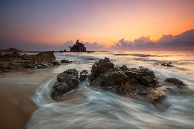 This depicts a serene sunset over a rocky beach with gentle waves flowing around the rocks. Ideal for travel websites, nature blogs, or advertisements promoting relaxation and beach destinations. Could also be used in environmental conservation campaigns or as a background image for inspirational quotes.