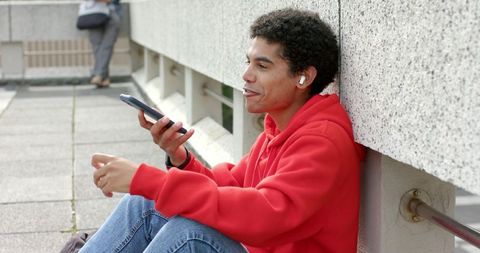 Asian man in red hoodie using smartphone and earbuds while leaning on urban parapet