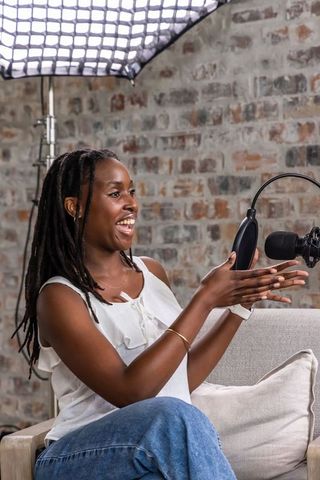 Recording podcast on sofa, woman hosting and gesturing toward microphone in studio