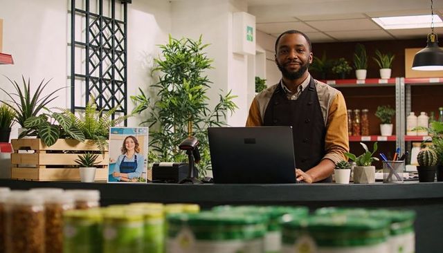 Smiling shop owner managing checkout with laptop and scanner in modern plant boutique