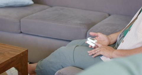 Mature woman sitting on sofa holding pulse oximeter monitoring health at home