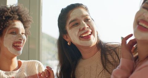 Diverse friends wearing facial masks laughing by sunny window in knitwear with gold hoops
