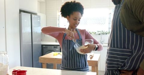 Couple cooking in bright kitchen mixing batter in glass bowl wearing striped aprons