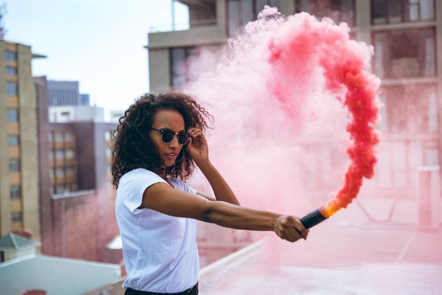 Front view of a hip young biracial woman wearing sunglasses and white t shirt, holding a smoke grenade with red smoke on an urban rooftop with building in the background