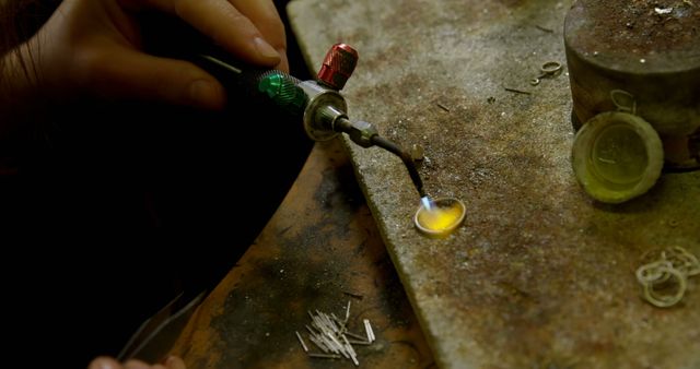 A person is melting a piece of metal using a small torch, with copy space. This activity is typical in jewelry making or metalworking, where precision heating is essential for shaping and soldering materials.