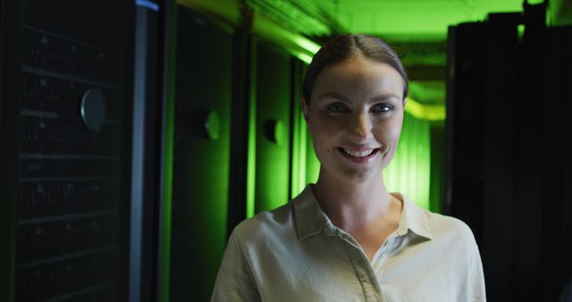 Image depicts a confident female IT engineer standing in a modern server room. She is smiling, wearing a light-colored blouse which contrasts with the green lighting in the background. This image can be used in various contexts such as technology articles, IT recruitment advertisements, cybersecurity campaigns, data center solutions, and promotional material for tech companies.