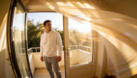 Morning breeze on balcony: man in white hoodie gazing through open glass doorway, golden light