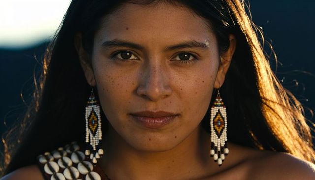 Sunlit woman facing camera, wearing beaded earrings and shell necklace, rimlit portrait