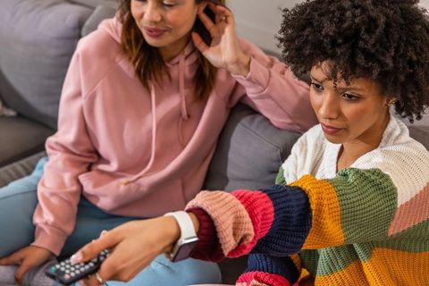 Diverse friends relaxing on sofa reaching for remote in striped sweaters and smartwatch