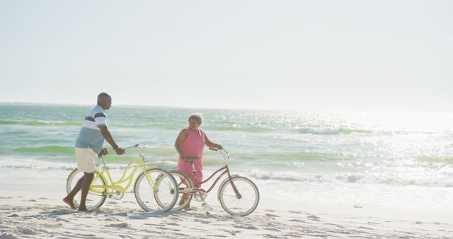 Senior couple walking their bicycles on a sunny beach. The ocean is calm and waves are gently hitting the shore. This can be used for promoting senior lifestyle, beach tourism, healthy outdoor activities, and travel guides.