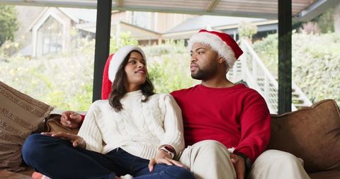 Couple sitting on couch wearing Santa hats and cozy sweaters sharing intimate holiday moment