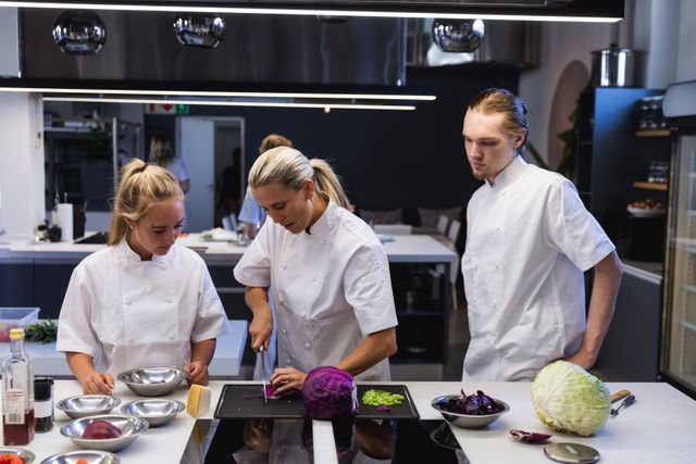 Group of chefs in a modern kitchen engaged in a cookery class. Female chef instructing two students, one male and one female, on slicing vegetables. Ideal for content related to culinary education, professional cooking, teamwork in the kitchen, and culinary workshops.